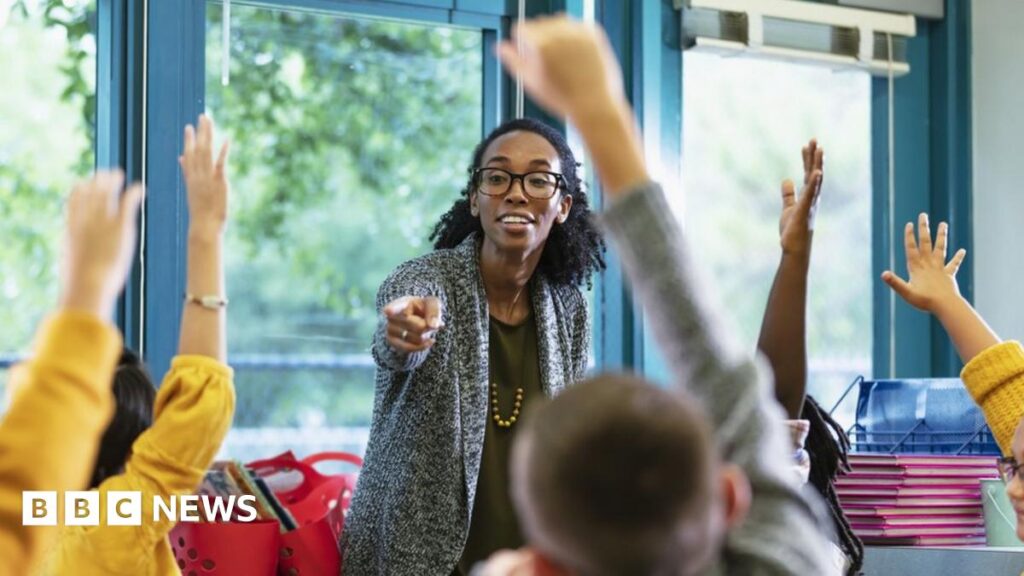 A teacher wearing black glasses and a grey cardigan smiles as she points towards a student with their hand in the air, surrounded by classmates also with their hands up.